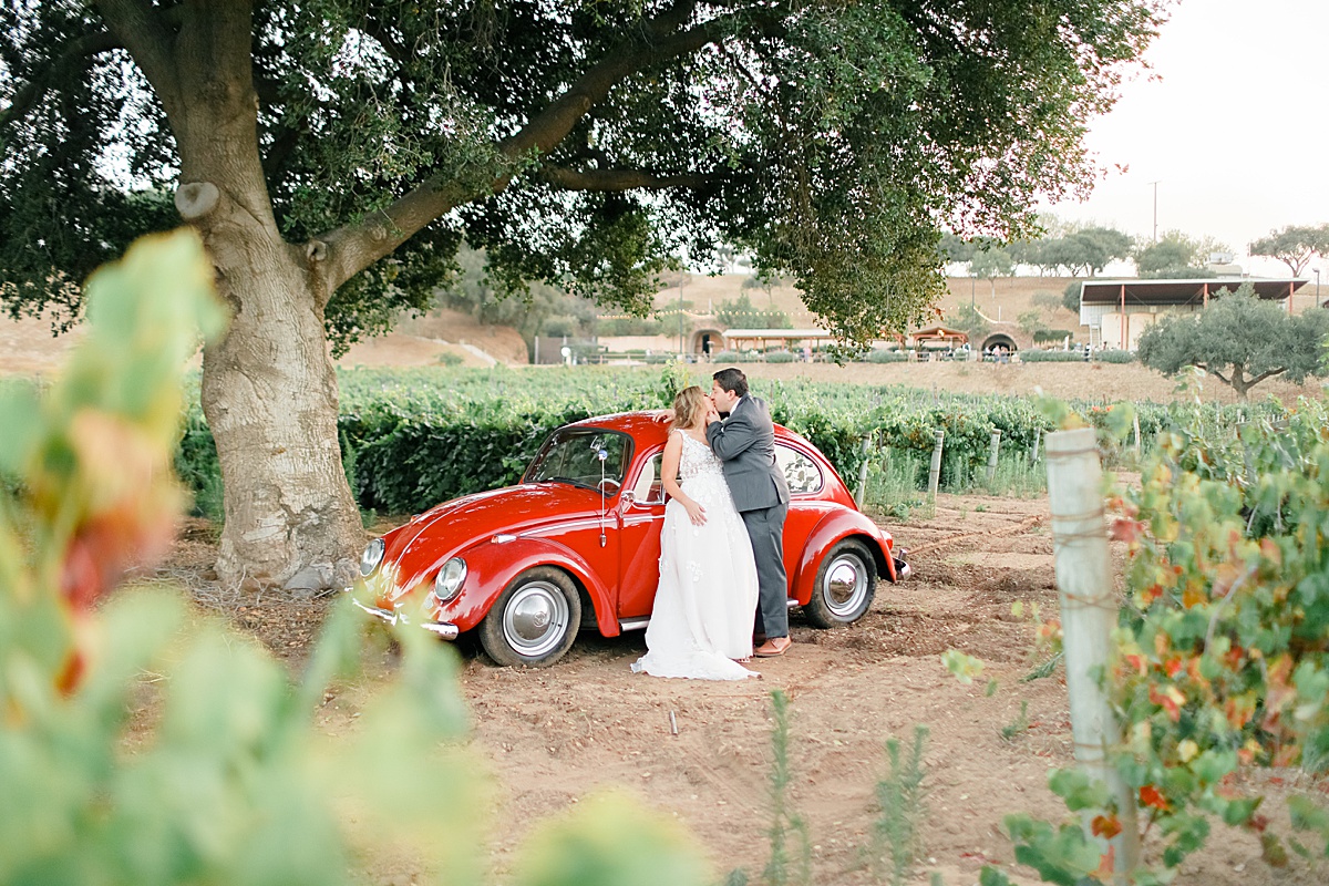 bride and groom kiss next to red volkswagen beetle in vineyard shot by destination wedding photographer
