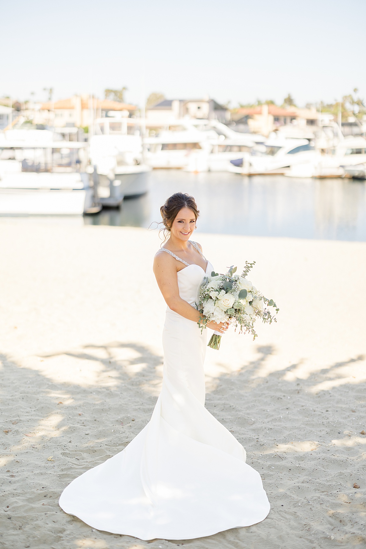bride in elegant minimalist gown poses with white rose bouquet on sandy ocean side before vow renewal at huntington beach
