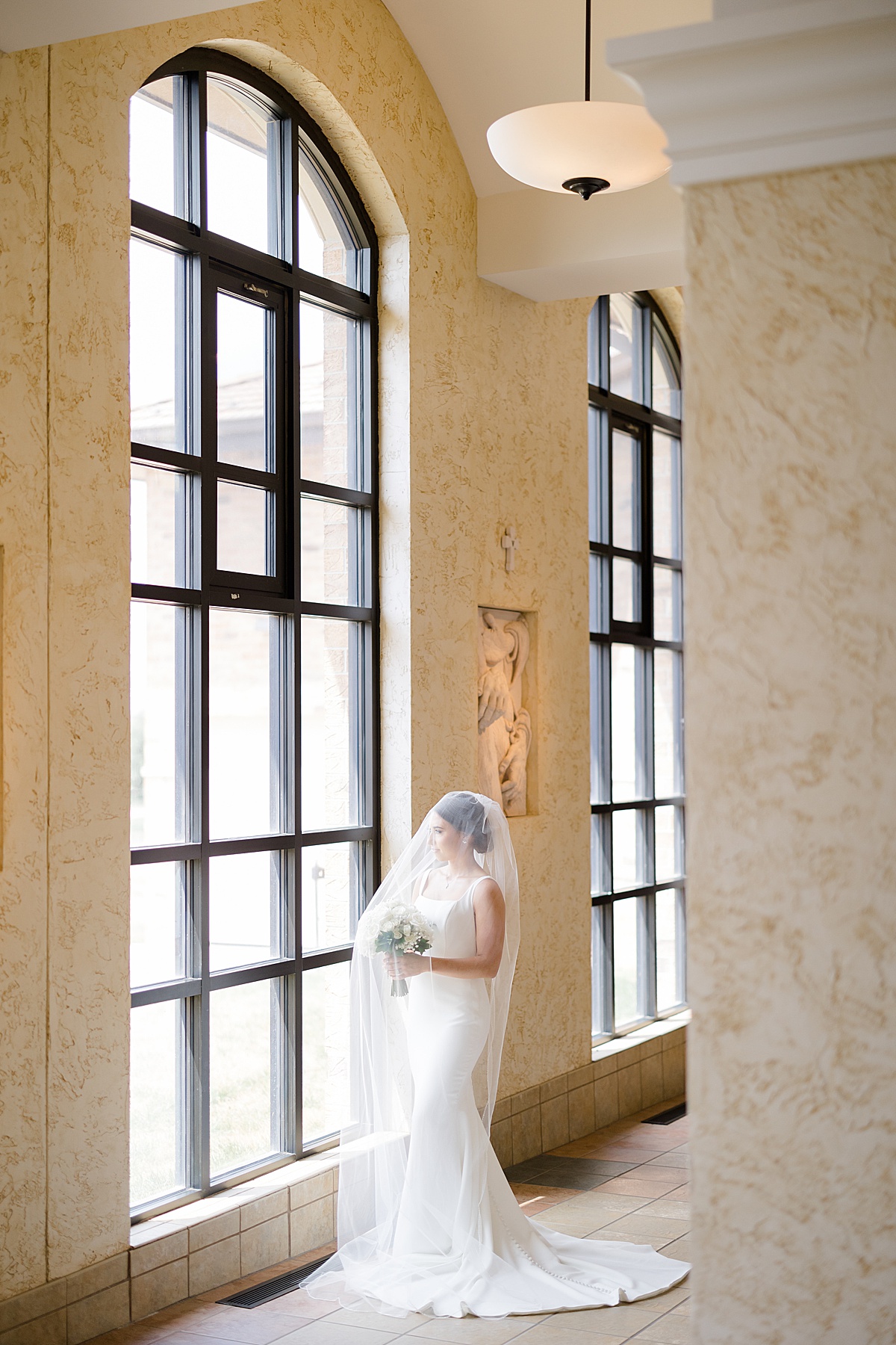 bride in minimalistic white gown poses by window before heartfelt Catholic ceremony