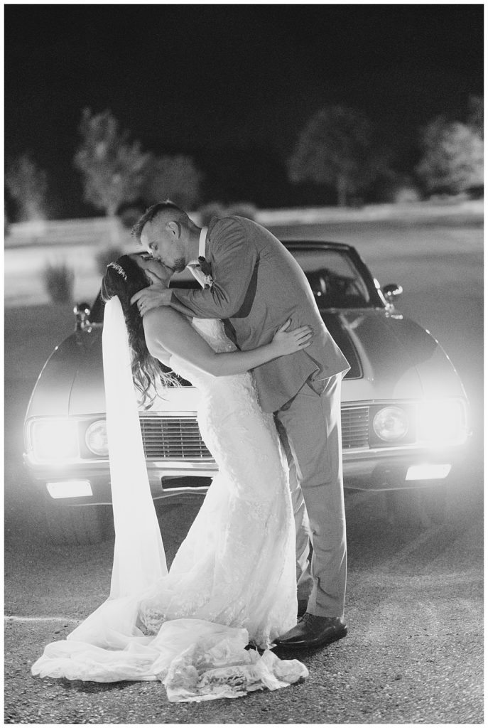 groom dips bride and kisses her in front of car at elegant Oklahoma wedding
